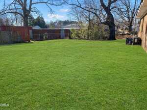 A large, well-maintained backyard with green grass, bordered by trees, a wooden fence, and a red shed, adjacent to a brick building. Some HVAC units are visible near the house. The sky is clear and blue.