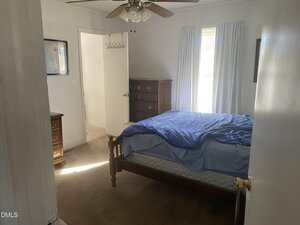 A small bedroom with a wooden bed frame, blue bedding, chest of drawers, and two windows with white curtains. A ceiling fan is above, and an open door reveals part of a bathroom.