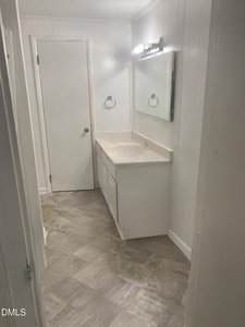 A small bathroom with white walls and a white vanity with a beige countertop under a rectangular mirror. The floor has a light patterned design, and there is a closed door at the end of the room.