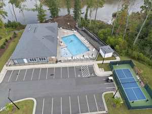 Aerial view of a recreational area with a rectangular swimming pool, poolside chairs, a small building, a parking lot with marked spaces including accessible spots, and a fenced tennis court surrounded by trees near a body of water.