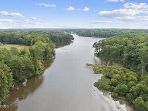 Aerial view of a wide river or lake with dense green trees on both sides under a blue sky with scattered clouds. The shoreline is irregular with a small sandy area visible in the foreground.