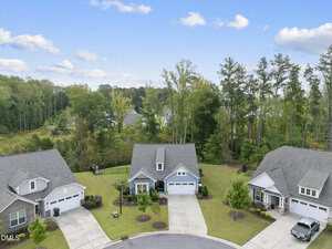 Aerial view of three single-family homes with gray roofs and driveways, arranged in a cul-de-sac. The houses have lawns and landscaping; behind them is a wooded area with tall trees and greenery.