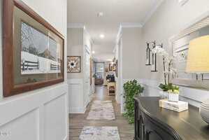 A hallway with light-colored walls and wainscoting, decorated with framed art, a mirror, plants, and a console table with a lamp and books. The hall leads to a living area with visible seating in the background.