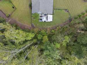 Aerial view of a house with a gray roof and fenced backyard, bordered by a grassy lawn. Dense green trees and forest run along the bottom edge of the image, separating the yard from the woods.