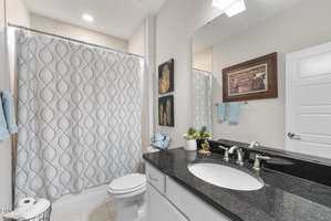 A bathroom with a white shower curtain featuring a blue pattern, a toilet, a black countertop with a sink, a mirror, wall art, and blue towels hanging on a rack.