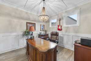 A home office with a wooden desk, brown leather chair, chandelier, table lamp, framed art, small decorative items, potted plant, printer, and a red electric guitar mounted on the wall near two windows.