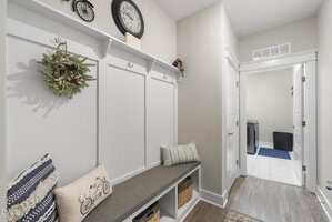 A mudroom with light-colored walls, a gray bench with cushions, baskets underneath, coat hooks above, a decorative wreath, a large clock, and an open doorway leading to a laundry room with washer and dryer.