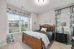 Bedroom with a wooden bed frame, gray bedding, black nightstand with lamp, two windows with floral curtains, a small white rocking chair, and carpeted floor. Walls are light-colored with artwork above the bed.