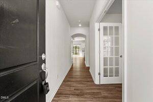 View from an open black front door into a hallway with wood flooring, white walls, a glass-paneled door to the right, and an archway leading to a bright room at the end.