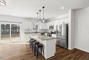 Modern kitchen with white cabinets, stainless steel appliances, a large granite island with four dark stools, pendant lights above, wood-look flooring, and sliding glass doors leading to a backyard.