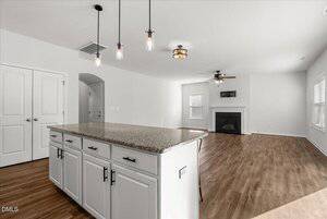 A modern open-plan kitchen and living area features a granite island with white cabinets, pendant lights, wood flooring, a ceiling fan, a fireplace, and large windows letting in natural light.