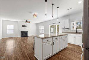 A modern open-plan kitchen and living area with white cabinets, granite countertops, wood flooring, pendant lights, a fireplace, and large windows letting in natural light.