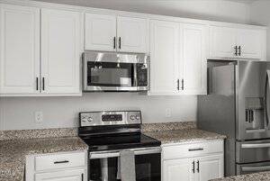 A modern kitchen with white cabinets, a stainless steel microwave above a matching oven, granite countertops, and a refrigerator to the right. A dish towel hangs from the oven handle.
