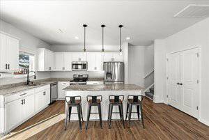 A modern kitchen with white cabinets, stainless steel appliances, granite countertops, three pendant lights, a large island with four black barstools, and wood-look flooring. A staircase and double doors are in the background.