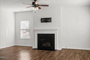 A bright, empty living room with light-colored walls, wood flooring, a black fireplace, a ceiling fan with lights, a small window with blinds, and a TV wall mount above the fireplace.