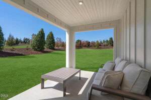 A covered patio with a beige outdoor sofa and a coffee table overlooks a well-maintained green lawn and landscaped garden with trees and shrubs under a clear blue sky.