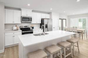 Modern kitchen with white cabinets, stainless steel appliances, a large island with a sink, and four beige cushioned stools. A dining table with six chairs is visible near sliding glass doors in the background.