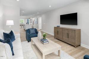 A modern living room with a white couch, blue armchair, and wooden coffee table topped with decor. A flat-screen TV is mounted above a wooden cabinet. The space features light wood floors and neutral walls.