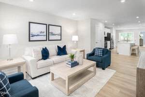 A bright, modern living room with a white sofa, two blue armchairs, a light wood coffee table, and two abstract paintings on the wall. The space opens to a kitchen and dining area with light wood flooring throughout.