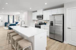 Modern kitchen with white cabinets, stainless steel appliances, and a center island with a sink and three beige cushioned stools. Part of a living room with blue accents is visible in the background.