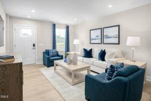 A living room with light wood floors, a white sofa, two blue armchairs, a wooden coffee table, and two table lamps. Blue curtains flank a window, and framed artwork hangs above the sofa. A door and console are on the left.