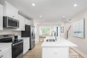 Modern kitchen with white cabinets, stainless steel appliances, and a large island with a built-in sink. Light wood flooring and neutral walls. Dining area and staircase are visible in the background.