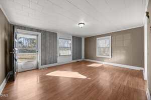 Empty living room with light brown laminate flooring, beige and gray walls, two large windows, a white ceiling with one light fixture, and a door with a window leading outside. Sunlight shines through the windows.