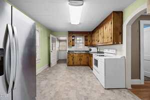 A kitchen with wood cabinets, white appliances including a stove and refrigerator, light green walls, a window, and a door leading outside. The floor has a gray patterned linoleum.