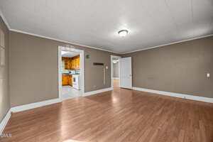 Empty room with wood laminate flooring, beige walls, white trim, and a white ceiling with a central light. Doorways lead to a kitchen with wood cabinets and another room with white walls and a window.