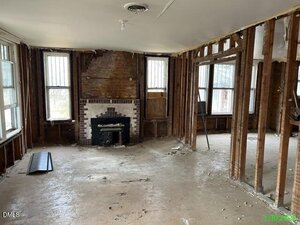 A stripped-down room with exposed wall studs, bare floors, and debris scattered throughout. A brick fireplace with a wood stove insert is centered on the back wall. The photo is dated 1/8/23.