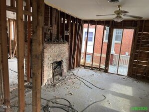 Room under renovation with exposed wall studs and wiring, debris on the floor, an unfinished fireplace, a ceiling fan, and large windows showing a brick building outside. Photo date is January 8, 2026.