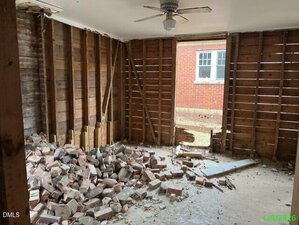 A room with exposed wall studs, a ceiling fan, and a large pile of broken bricks and debris on the floor. A window provides a view of an exterior red brick wall.