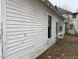 The side of a white, weathered wooden house with visible dirt and peeling paint, a few windows, and an overgrown yard with dry grass. Another house is partially visible in the background.