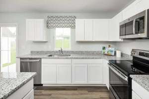 A modern kitchen with white cabinets, granite countertops, stainless steel appliances, a window above the sink, wood flooring, and a sliding glass door to the left. A small cookbook and utensils are on the counter.