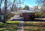 A small, single-story brick house at 1324 Raleigh Road in Oxford with a covered front porch, white railing, and a concrete walkway leading to the door. The yard has patchy grass, leafless trees, and some bushes under a clear blue sky.
