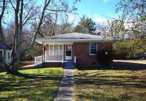 A small, single-story brick house at 1324 Raleigh Road in Oxford with a covered front porch, white railing, and a concrete walkway leading to the door. The yard has patchy grass, leafless trees, and some bushes under a clear blue sky.