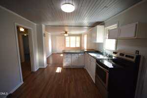A kitchen with white cabinets, stainless steel appliances, and a double sink, opening into a dining area with wood-style floors, gray walls, and a ceiling fan. Natural light comes in through several windows.