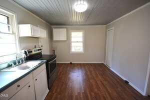 A small kitchen with wood-look flooring, white cabinets, a stainless steel stove, a stainless steel sink under a window, and light gray walls with a gray plank ceiling. A single ceiling light is on.