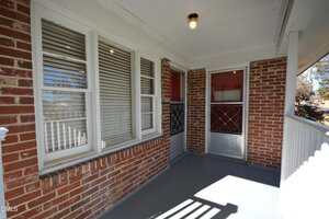A covered front porch with red brick walls, a gray painted floor, white trim, three windows with blinds, a light fixture on the ceiling, and a red and silver screen door.
