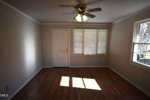 A small empty room with light brown wood flooring, beige walls, a white front door, a window with blinds, a side window, and a ceiling fan with lights turned on. Sunlight is shining through the windows onto the floor.
