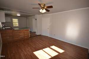 A mostly empty room with wood laminate flooring, white walls, a ceiling fan with lights, and a partial view of a kitchen area with a wood-paneled counter and white cabinets. Sunlight enters through a window.