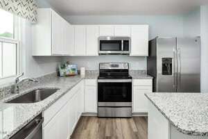 Modern kitchen with white cabinets, stainless steel appliances including a refrigerator, oven, and microwave, granite countertops, a double sink, and light blue walls. Wood-look flooring is visible.