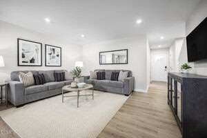 A modern living room with two gray sofas, a round wooden coffee table, and a light rug on wood flooring. Black and white artwork decorates the walls, and a TV sits on a console opposite the couches.