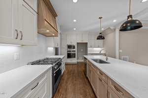 Modern kitchen with white cabinets, stainless steel appliances, a gas stove, built-in oven, white subway tile backsplash, center island with sink, pendant lights, and medium tone wood flooring.