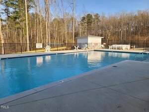 An outdoor swimming pool with clear blue water, surrounded by a concrete deck, white pool chairs, and a shed. Leafless trees and a black metal fence border the pool area under a clear sky.