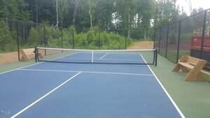 A blue and green outdoor tennis court surrounded by a black chain-link fence, with two wooden benches on either side and dense green trees in the background.