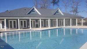 A large outdoor swimming pool in front of a gray building with white trim, multiple glass doors, dormer windows, and a covered patio area. Tall trees are visible in the background under clear skies.