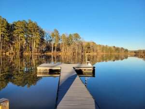A wooden dock extends over a calm, reflective lake bordered by tall trees under a clear blue sky, with the shoreline and trees mirrored in the still water.