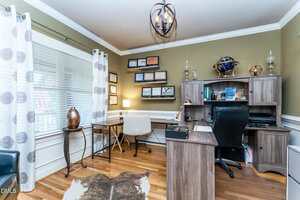 A home office with a wooden desk, shelving, a globe, and various certificates on the wall. A window with patterned curtains lets in light, and a white chair sits at a smaller desk by the window. Hardwood floor with a cowhide rug.