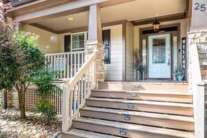 Front porch of a beige house with stone accents, white railing, wooden steps with built-in lights, a welcome sign, potted plants, and a glass front door with sidelights and a decorative wreath.